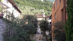 A stone pathway leading through a courtyard with buildings on either side, surrounded by greenery and mountains in the background. - Great Price... Romantic Apartment Near Beaches and Nightlife (Malcesine)