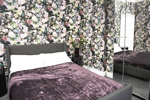 A bedroom with a floral wallpaper, a bed with a purple bedspread, a nightstand, a mirror, and a dresser. - Fabulous & Funky 3 Bedroom Flat in East London. Close to Underground & 24h Tesco (London)