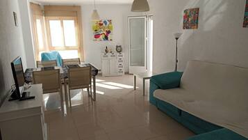 Una sala de estar moderna con un sofá color turquesa, un área de comedor con mesa y sillas, un armario blanco y un amplio ventanal con cortinas.