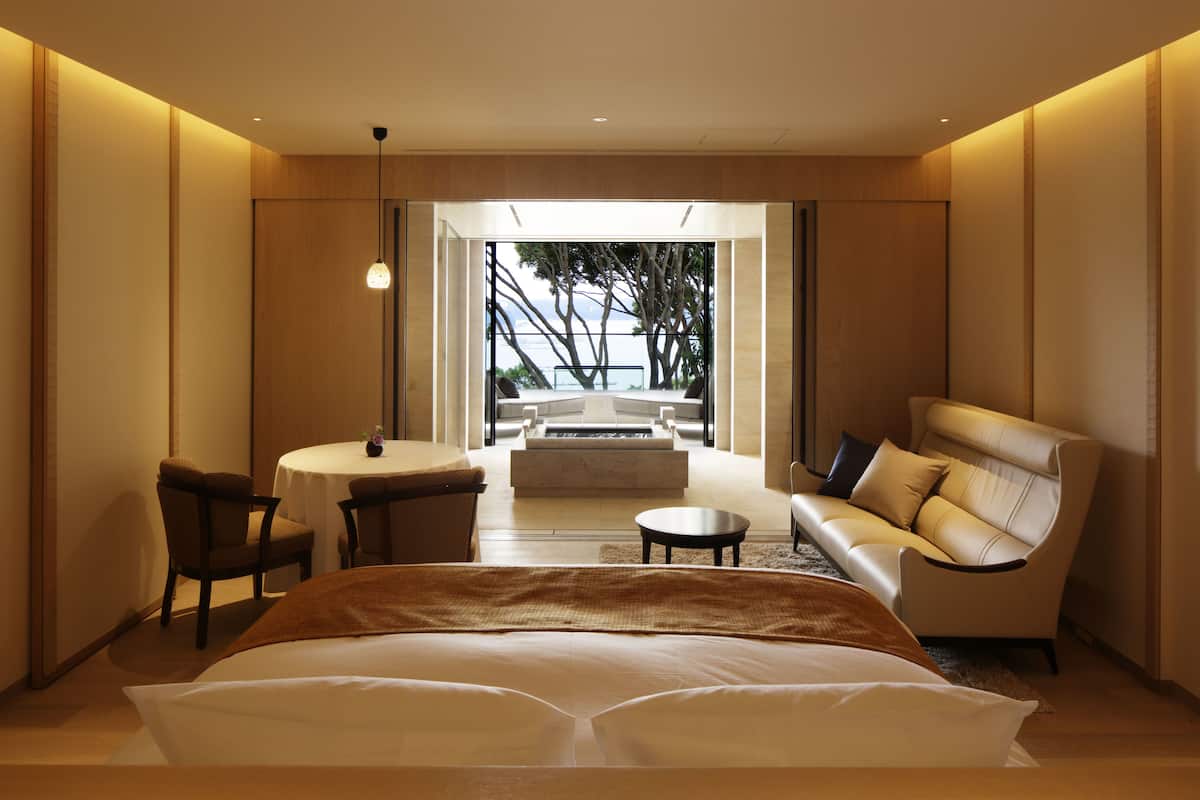 A modern hotel room with a large bed, a sofa, a dining table, and a view of the outdoors.