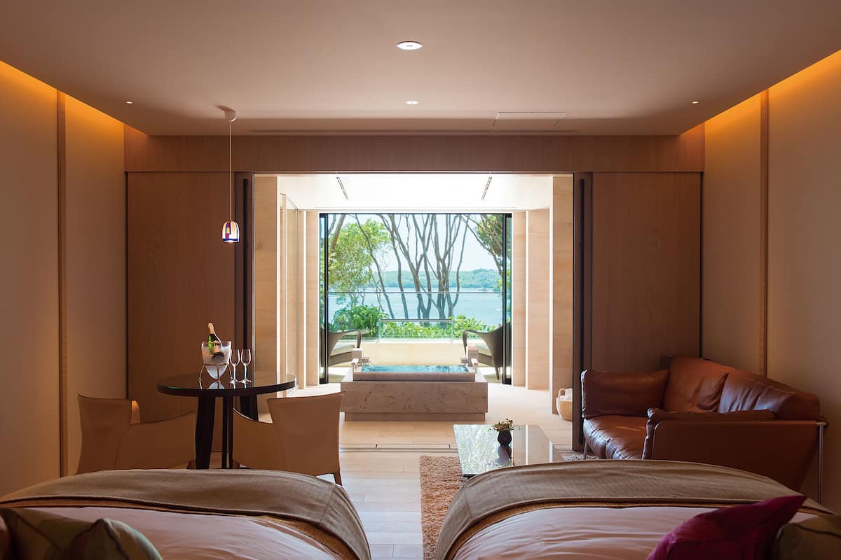 A modern hotel room with two beds, a sitting area with a view of the sea, and a small table with a bottle and glasses.