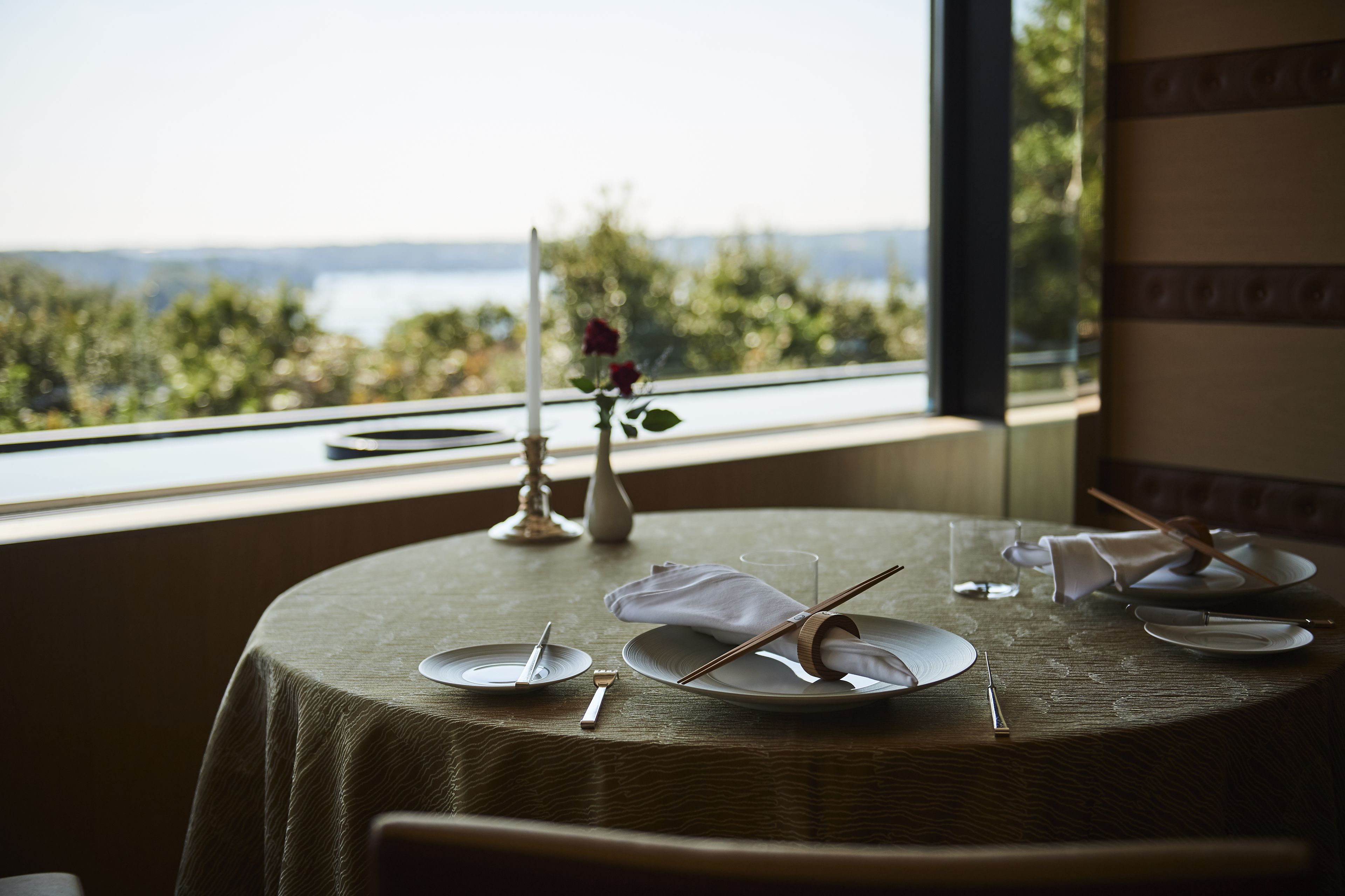 Mesa de comedor dispuesta con platos, cubiertos y un florero con una sola rosa, con vistas a un paisaje pintoresco.