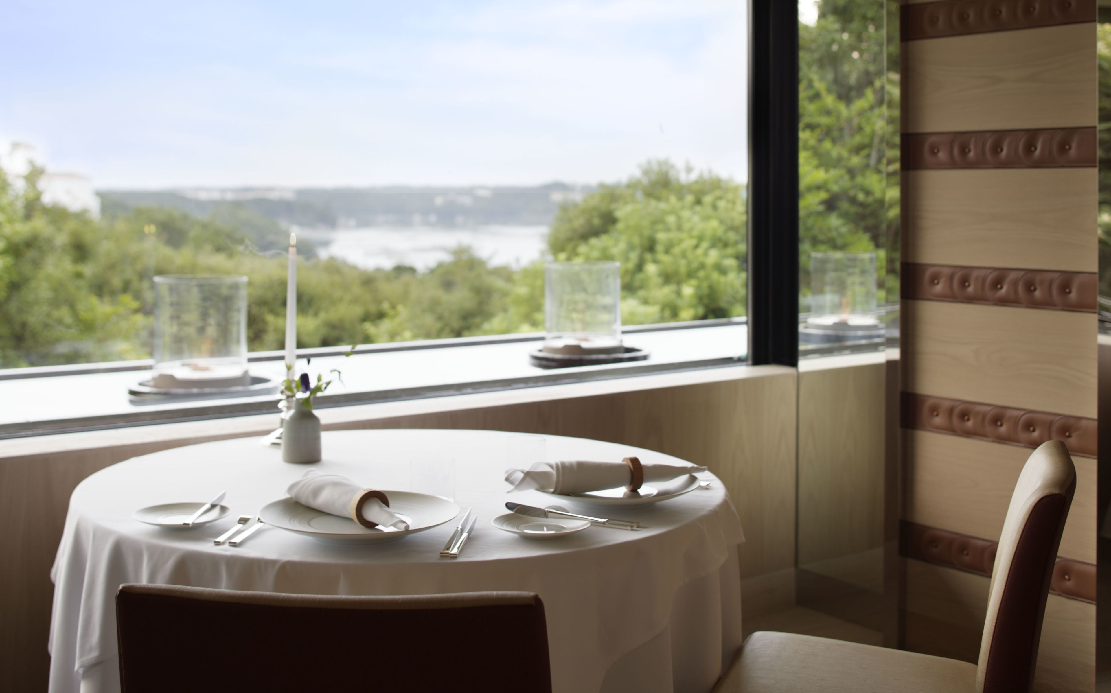 Mesa de comedor preparada para cuatro con vista a una masa de agua y vegetación a través de una ventana.