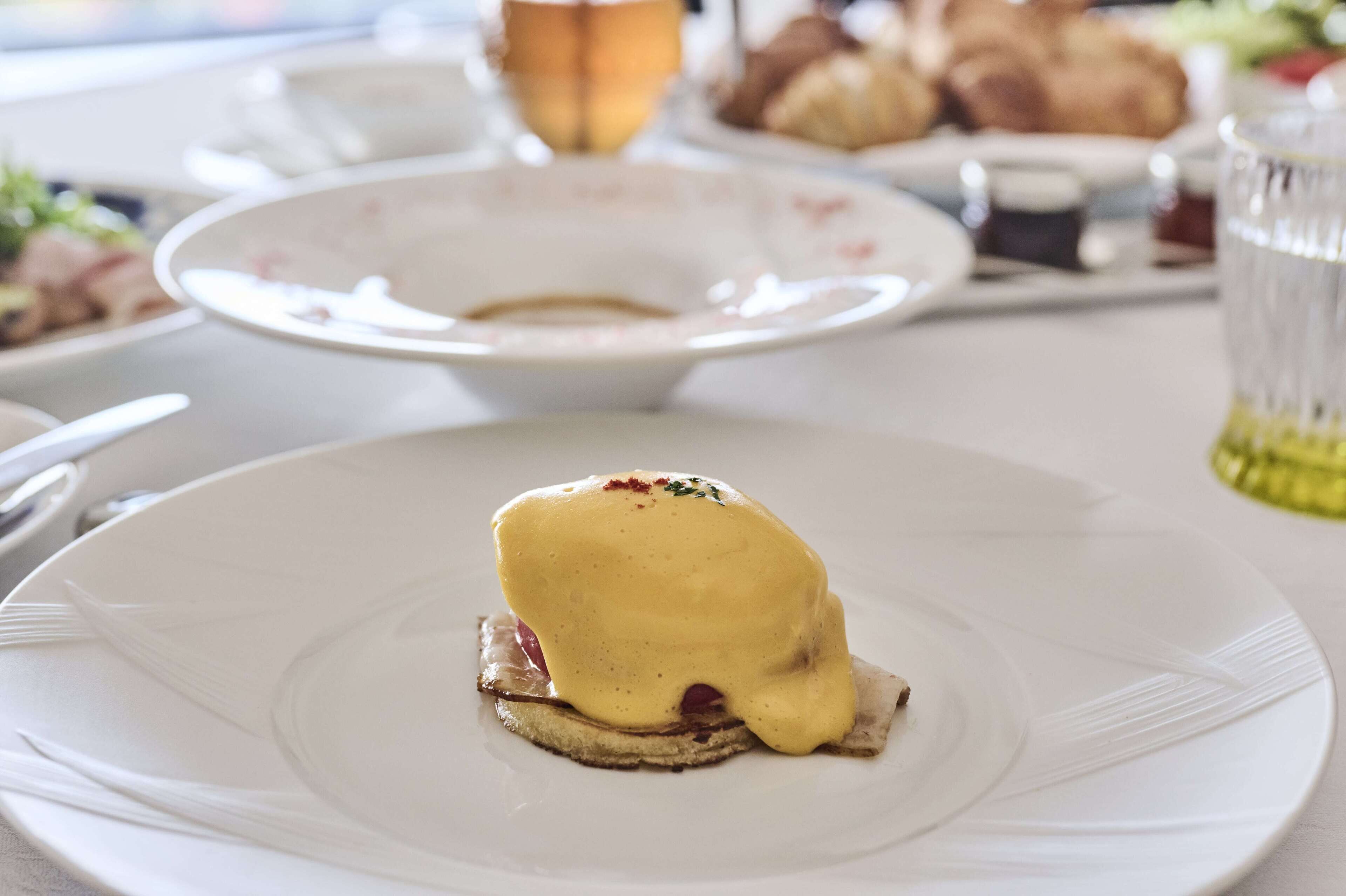 Een ontbijttafel met eieren benedict en een schaal fruit op de achtergrond.