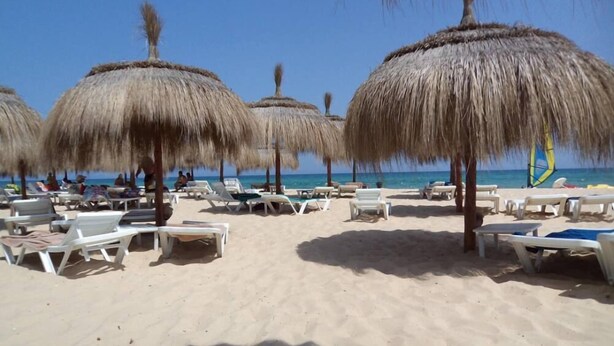 Una playa con sombrillas de paja y sillas reclinables blancas.