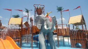 Un paseo en bote con forma de elefante, con un jinete, rodeado de construcciones de madera y palmeras.