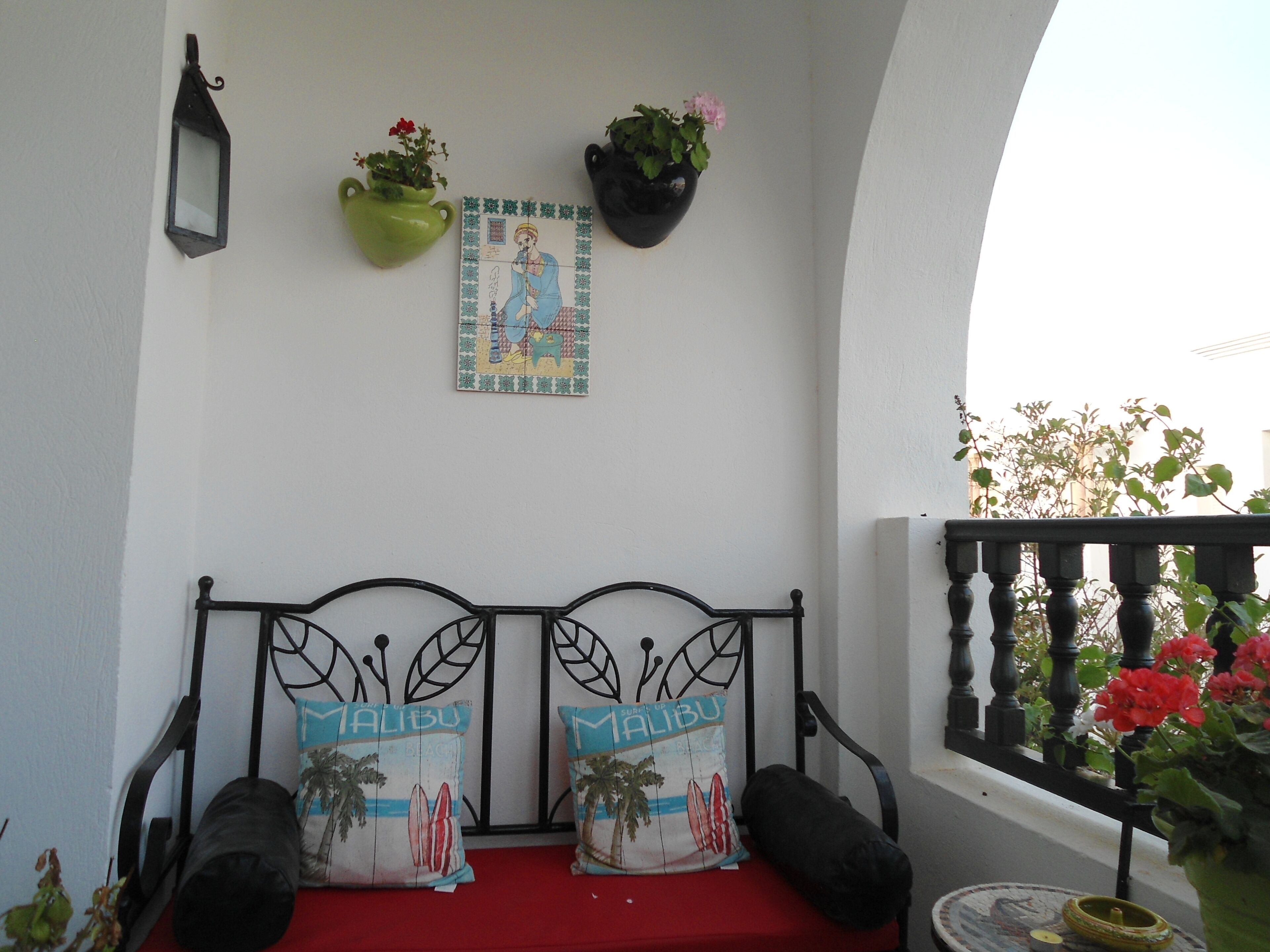Een balkon met een rode, gewatteerde bank, twee kussens met de tekst 'Malibu', een aan de muur bevestigde lantaarn en hangende plantenbakken met bloemen.