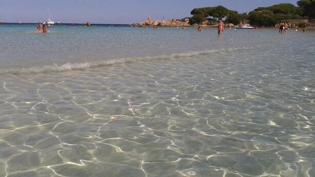 Ein Strand mit klarem, seichten Wasser und Felsformationen im Hintergrund.
