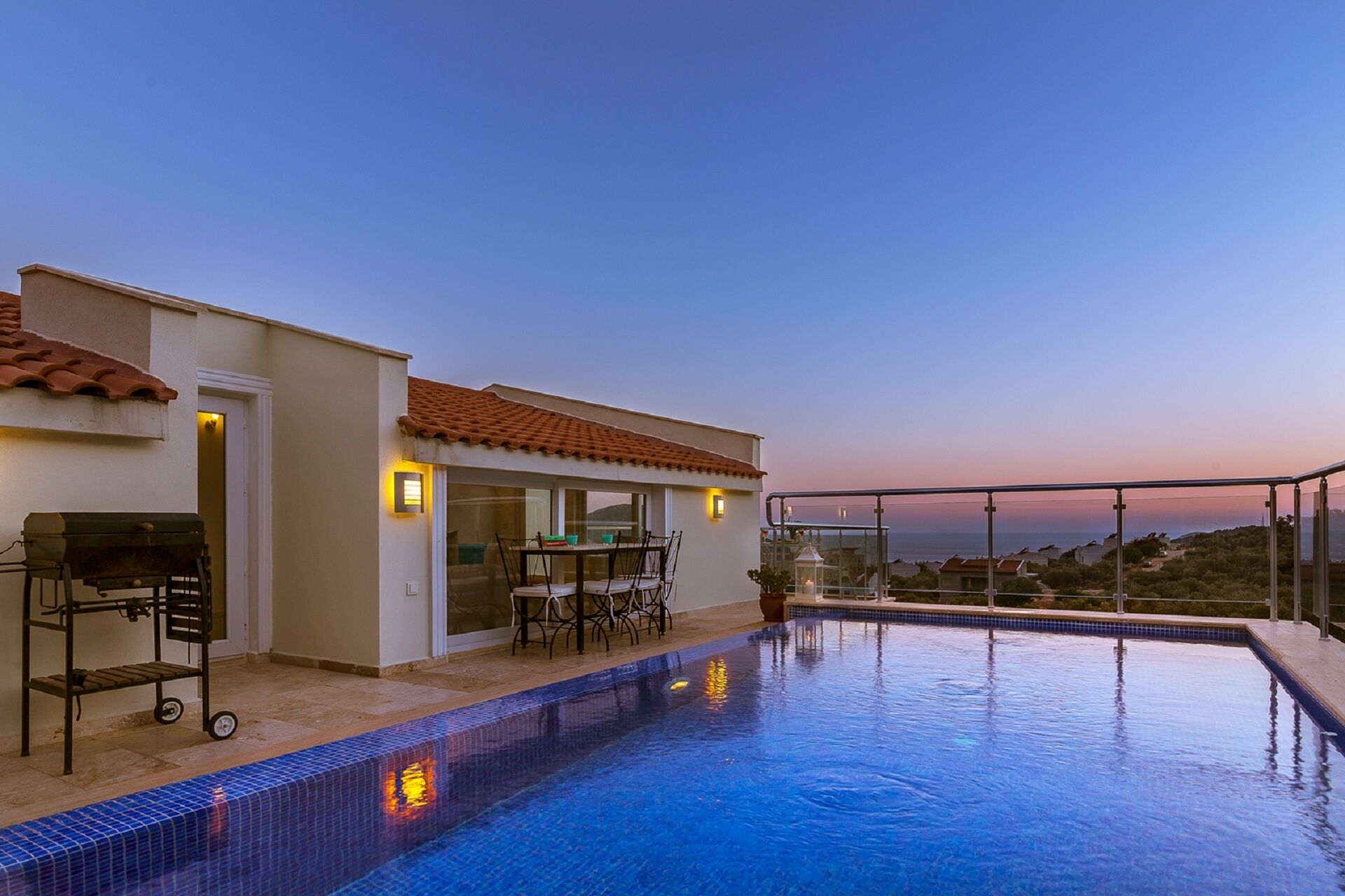 A swimming pool with a tiled edge, a barbecue grill, and a table with chairs outside a building with a tiled roof.