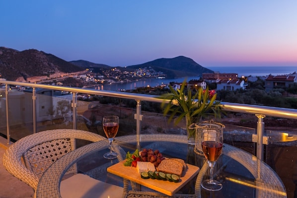 A balcony with wicker chairs, a glass table, a tray with cheese, grapes, cucumber slices, and two glasses of wine.