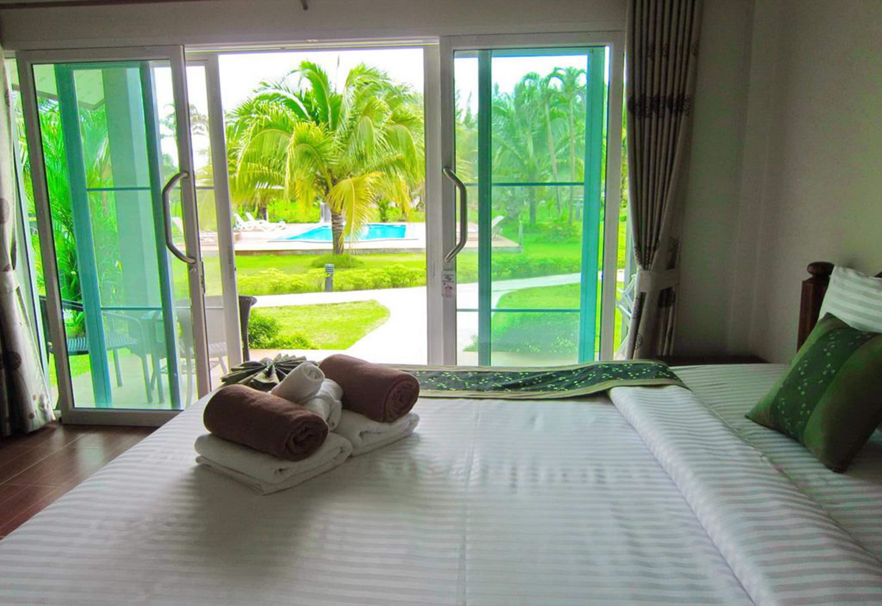 A hotel room with a bed, a view of a pool and palm trees through a sliding glass door, and a set of towels on the bed.