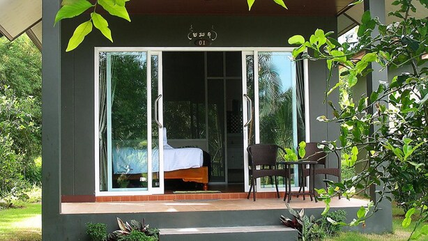 Un bâtiment moderne avec une entrée en verre, un lit visible à l’intérieur et un petit espace extérieur aménagé avec des chaises et une table.