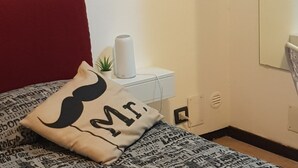 Un dormitorio con cabecera roja, edredón estampado, almohada con diseño de bigote, mesita de noche blanca con rollo de papel higiénico, una planta pequeña y un espejo en la pared.