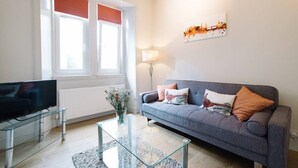Una sala de estar moderna con un sofá gris, un televisor de pantalla plana, una mesa de centro de vidrio y una pintura de un paisaje urbano en la pared.