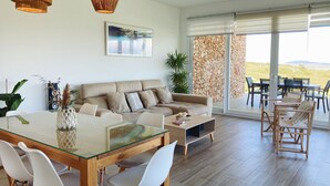 A modern living room with a glass dining table, beige sofa, and a large window with a view of the countryside.