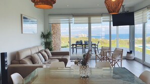 A modern living room with a glass dining table, comfortable seating, and a large window offering a scenic view.