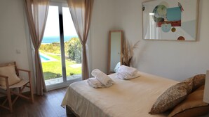 A bedroom with a bed, a chair, a large window with curtains, and a painting on the wall.