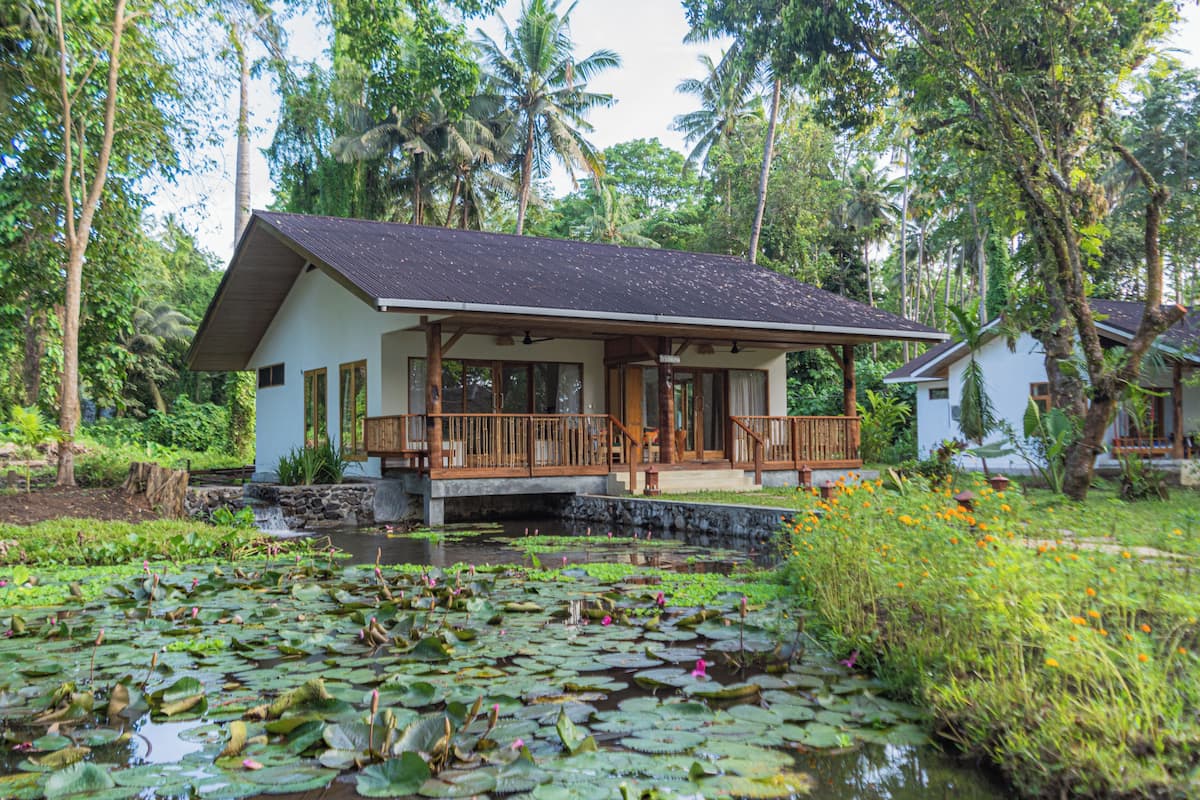 A bungalow with a porch, surrounded by a pond with lily pads and water lilies, and tropical vegetation in the background.