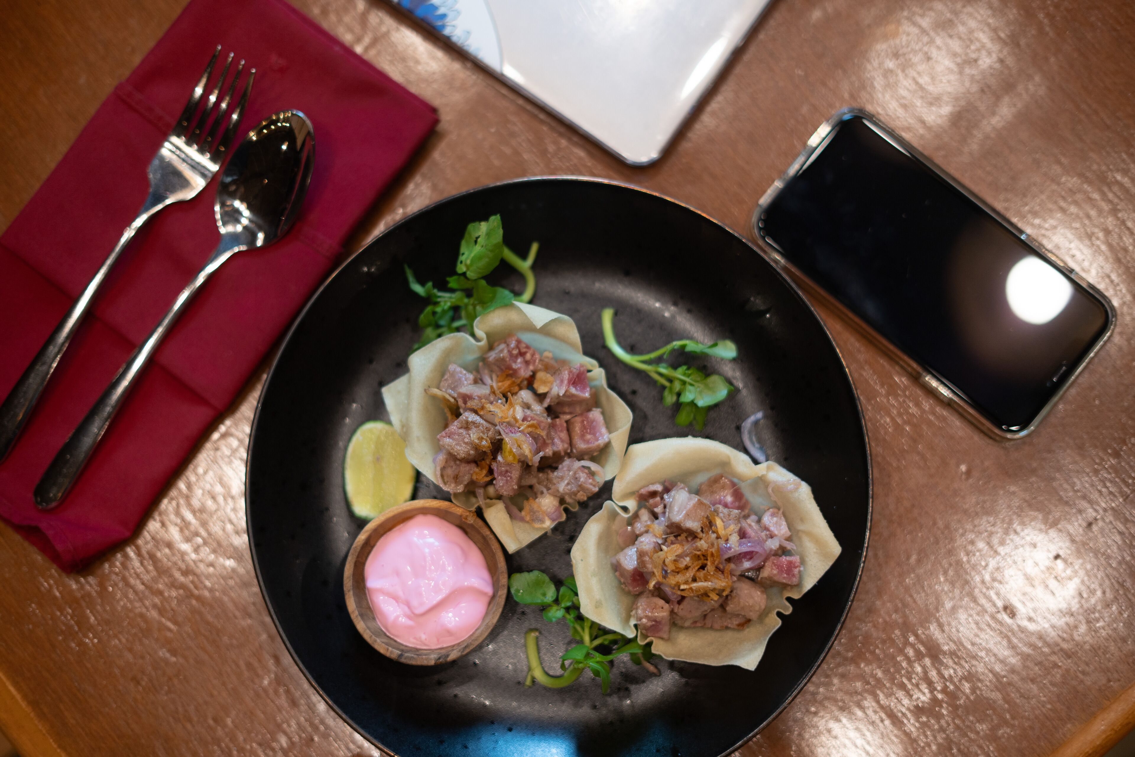 Un plato de comida con un recipiente pequeño de salsa, un tenedor, una cuchara sobre una servilleta y un teléfono inteligente en una mesa.