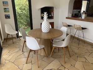A round wooden table with a pineapple centerpiece, surrounded by white chairs with wooden legs, in a room with a stone floor and a view of greenery outside.