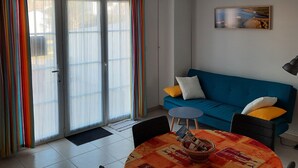 Een woonkamer met een blauwe bank, kleurrijke gordijnen, een eettafel met een tafelkleed met dessin en een schilderij aan de muur.