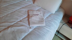 Een net opgemaakt bed met een witte deken en kussens, een gevouwen handdoek met de tekst 'La Cezarine Boutique' en een nachtkastje met een lamp.