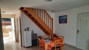 Een eetgedeelte met een houten tafel en stoelen, een kleine keuken met een koelkast en een spoelbak, en een trap die naar een bovenverdieping leidt.