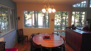 A dining area with a round wooden table, six chairs, a chandelier, and large windows with a view of trees. - Neil Bay Waterfront Family And Pet Friendly Property!<br>PCUP00-13-0010  (Roche Harbor)