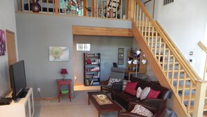 A living room with a brown leather sofa, a small coffee table, a bookshelf, a TV on a stand, and a staircase. - Neil Bay Waterfront Family And Pet Friendly Property!<br>PCUP00-13-0010  (Roche Harbor)