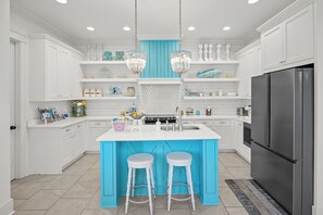 A modern kitchen with white cabinets, a blue accent wall, two bar stools, a stainless steel refrigerator, and pendant lights.