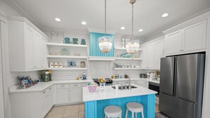 Cocina moderna con armarios blancos, una isla azul, dos taburetes de bar, un refrigerador de acero inoxidable y adornos en estantes.