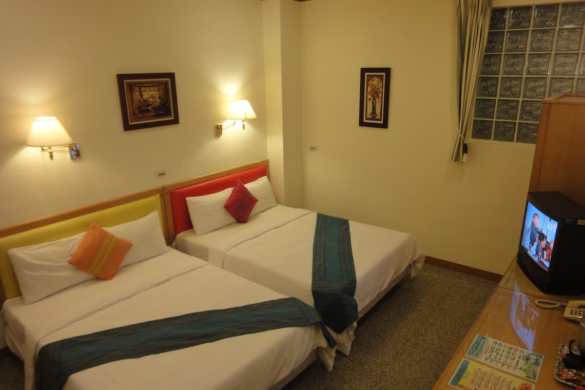 A hotel room with two beds, each with a pillow and a blanket, a TV on a wooden table, and wall-mounted lamps.
