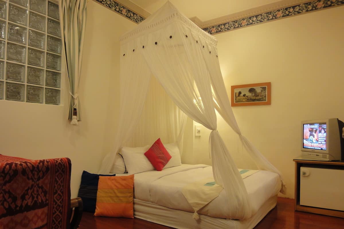 A bedroom with a bed, a television, a window with curtains, and a patterned sofa.