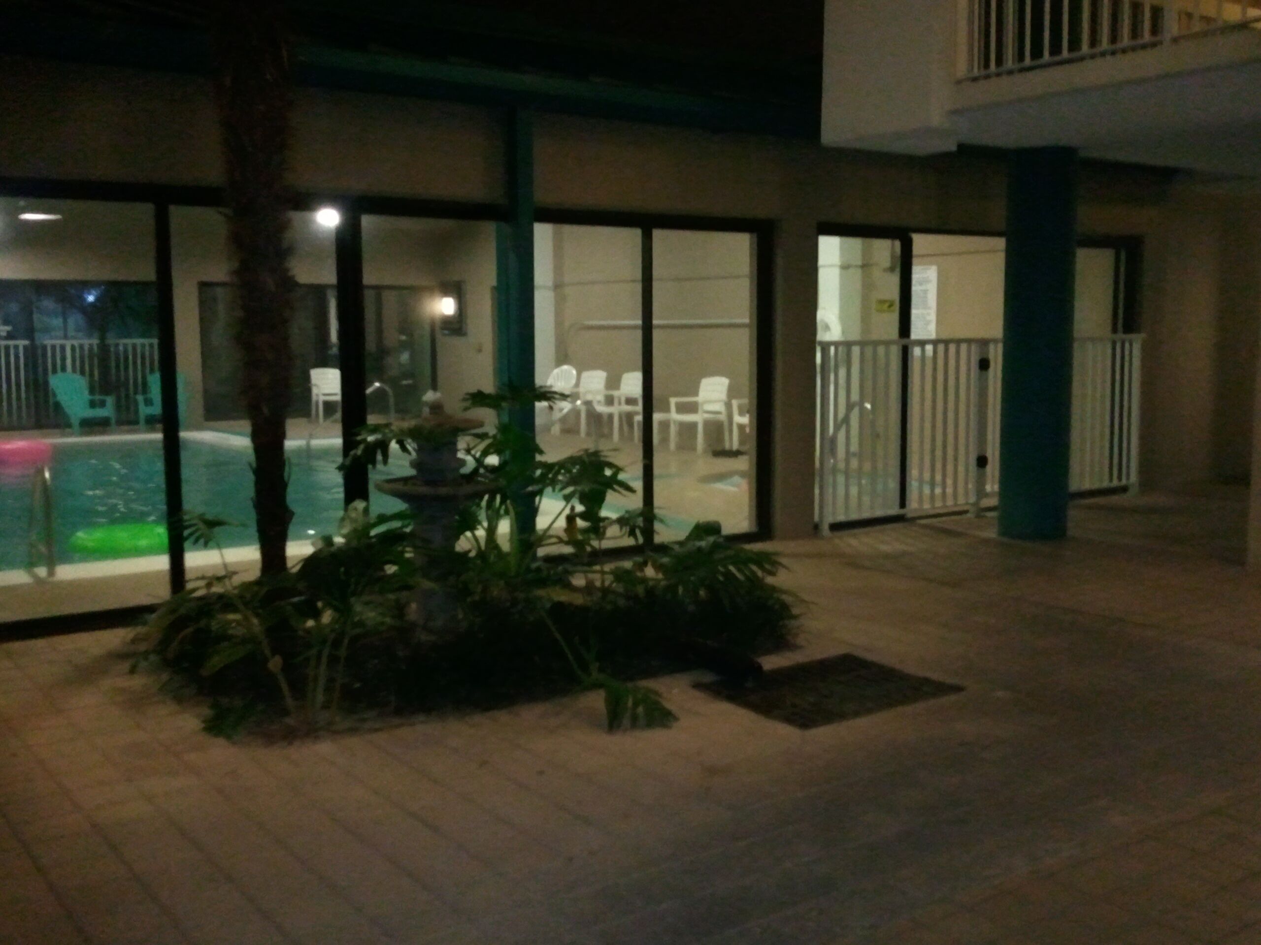 An outdoor pool area with a fountain, surrounded by potted plants and white chairs.