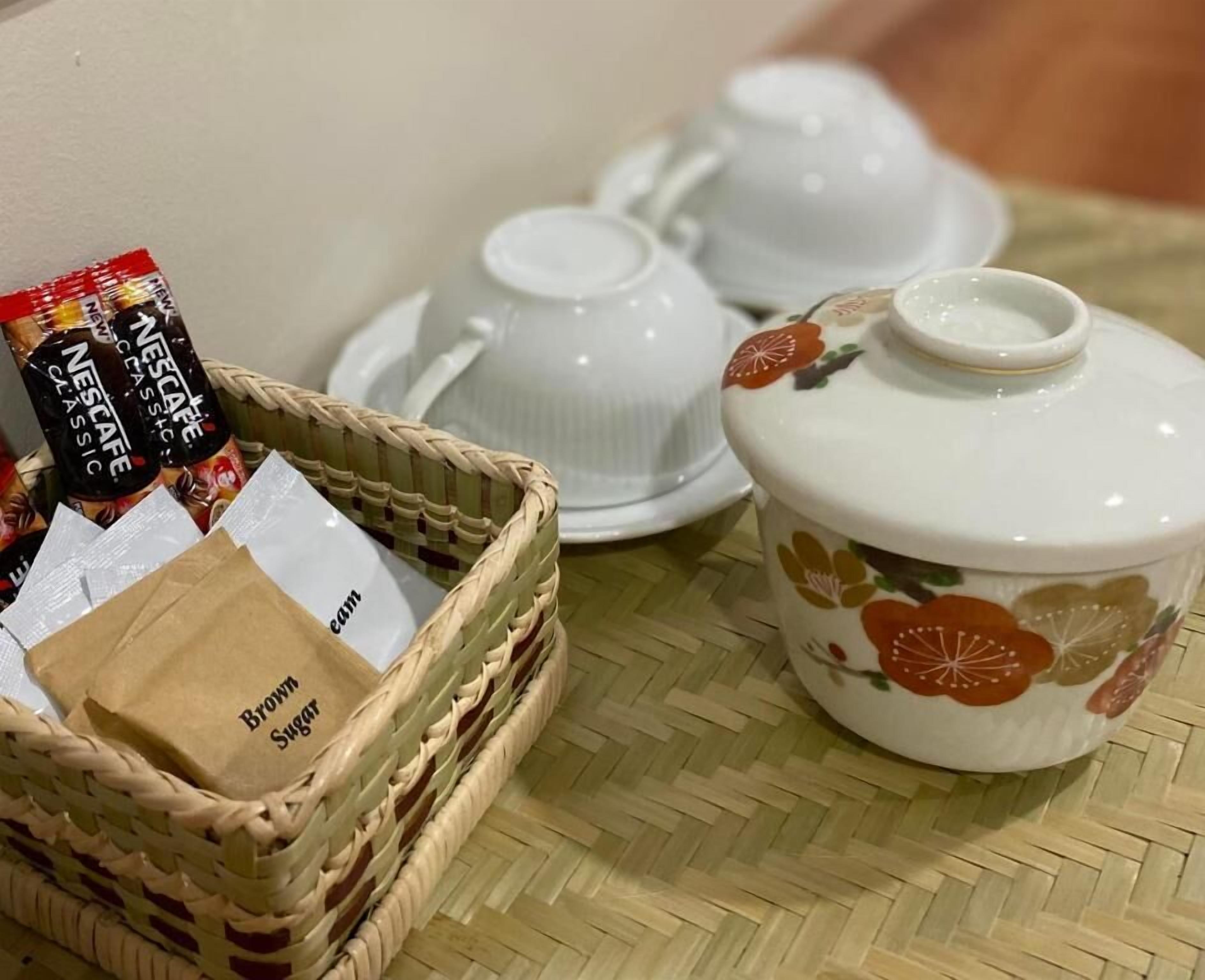 Una cesta de mimbre con una bolsa de papel marrón etiquetada 'Brown Sugar' y sobres de café Nescafe Classic, junto a una tetera blanca con diseño floral y tazas a juego sobre un tapete tejido.