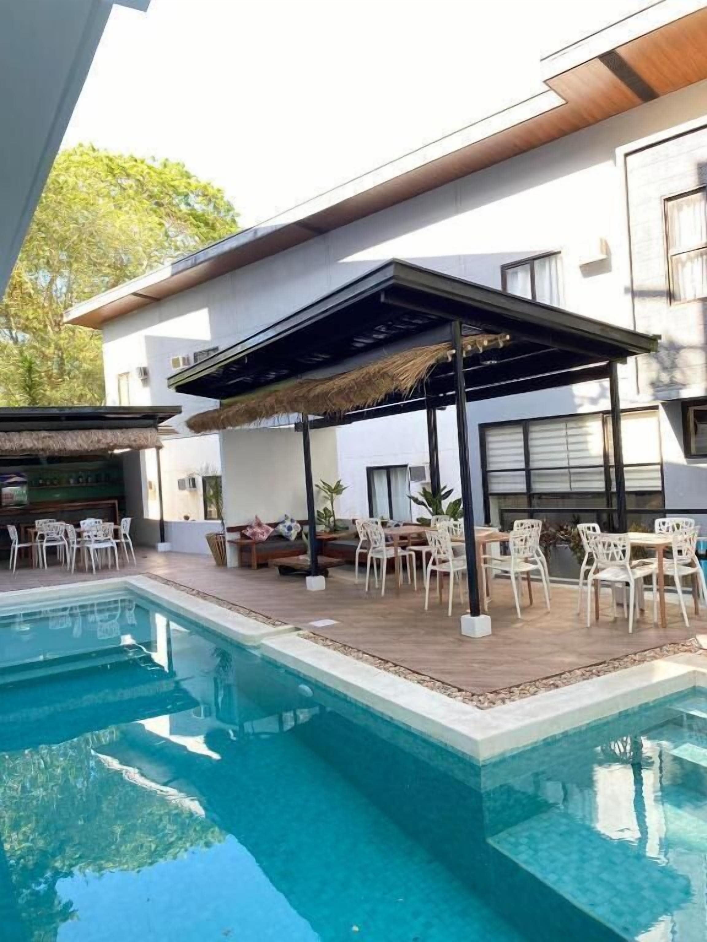 Un área de piscina moderna al aire libre con zona de estar techada, sillas de descanso y un bar.