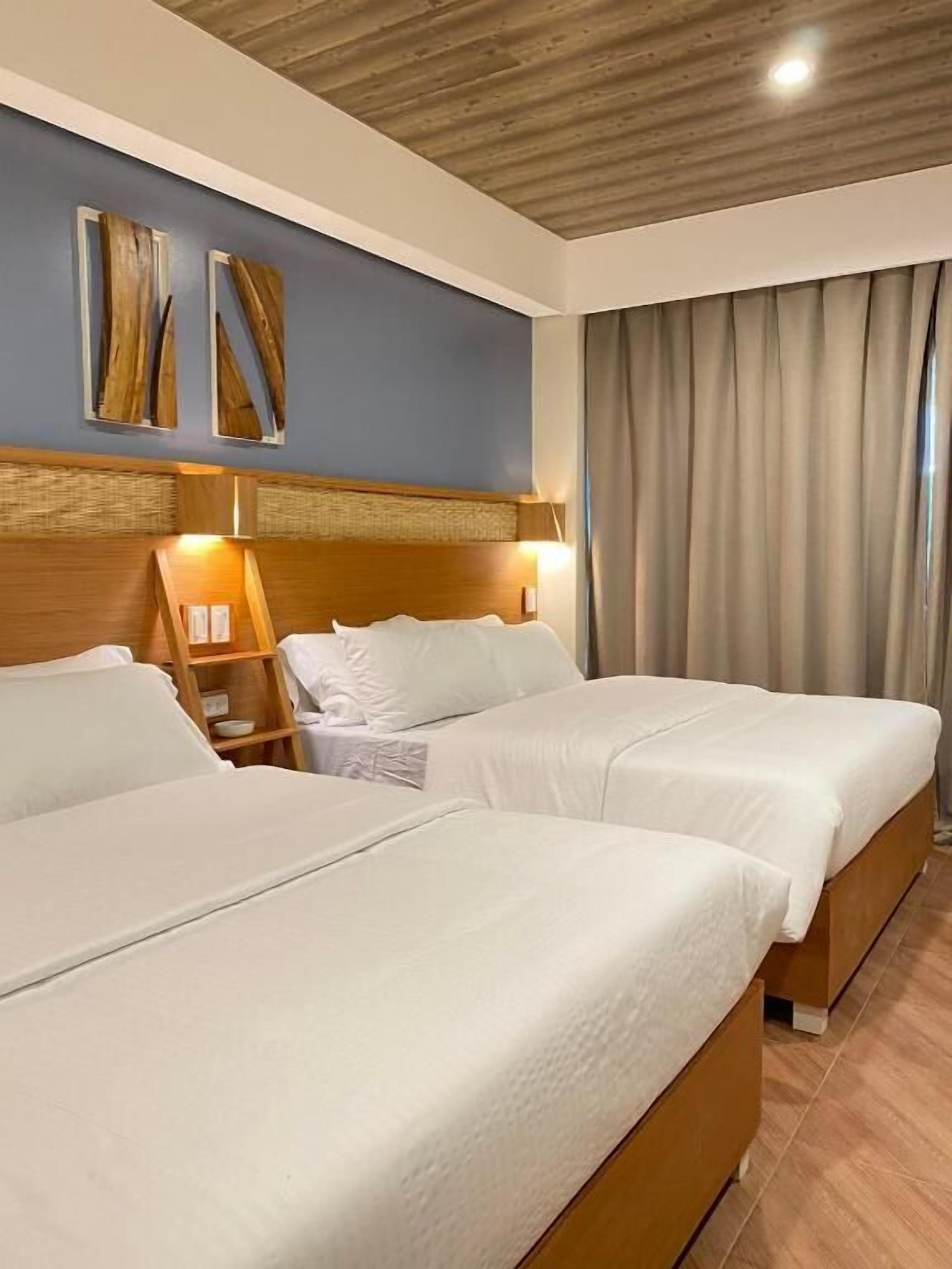 Een hotelkamer met twee bedden, een houten hoofdbord, een nachtkastje en een groot raam met gordijnen.