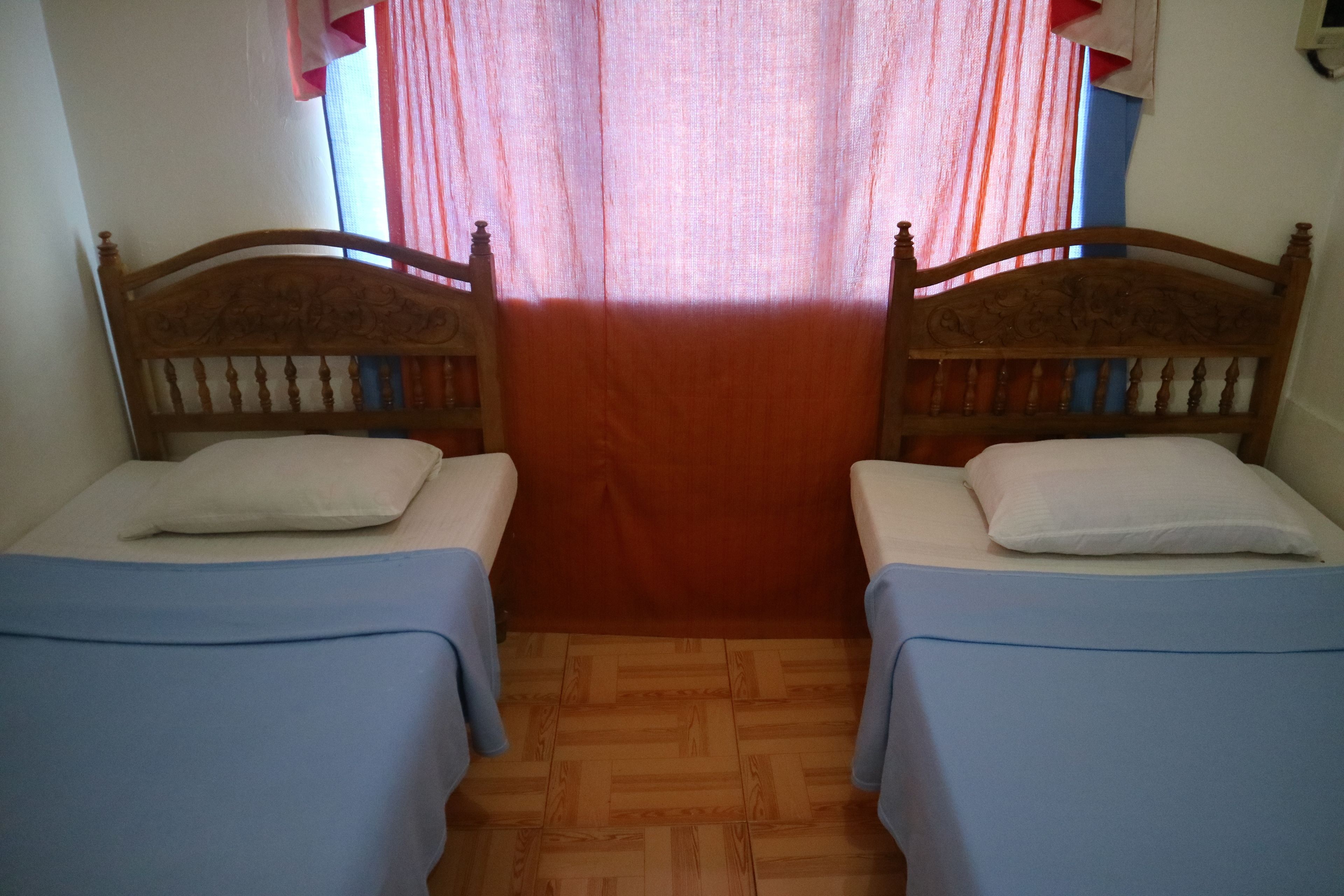 A room with two wooden beds, each with a blue bedspread and white pillows. The room has a wooden floor and a window with red curtains.