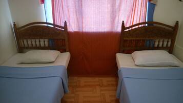 A room with two wooden beds, each with a blue bedspread and white pillows. The room has a wooden floor and a window with red curtains.