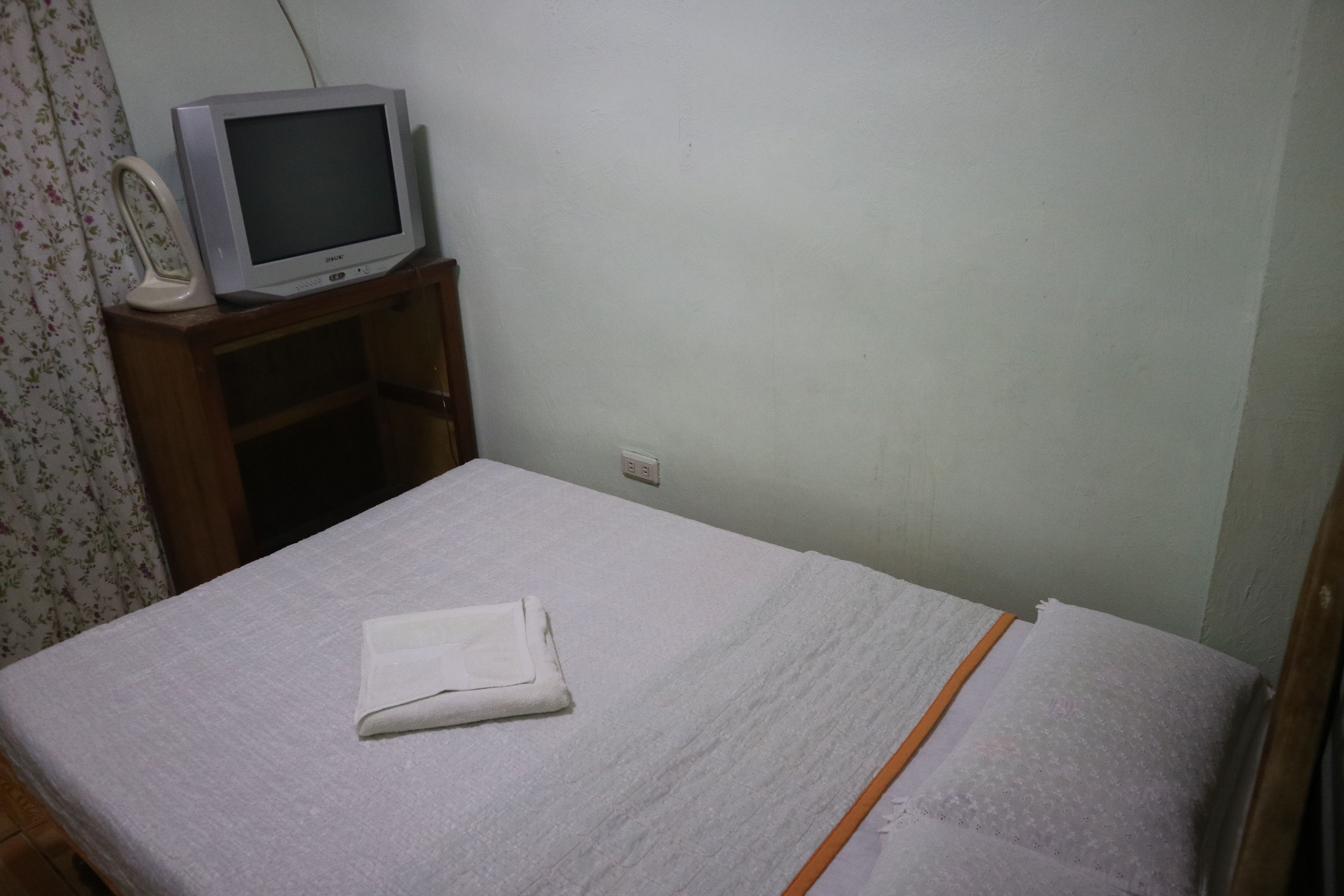 A simple room with a bed, a television on a wooden stand, and a power outlet on the wall.