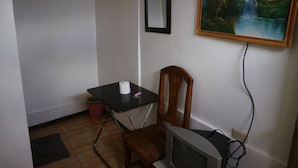 Een kleine kamer met een houten stoel, een bureau met een computer, een televisie en een schilderij aan de muur.
