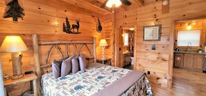 A wooden cabin interior with a bed, two lamps, a wall decal of a deer and trees, a framed picture, and a kitchen area.