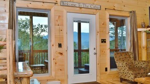 Ein Interieur einer Holzhütte mit einer Holztür, einer Bank, einem Stuhl und Blick ins Freie.