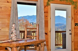 A wooden cabin interior with a dining table, a Christmas tree, and a view of mountains through large windows.