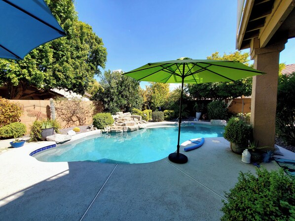 Une piscine avec un parasol vert, entourée de rochers et de verdure.