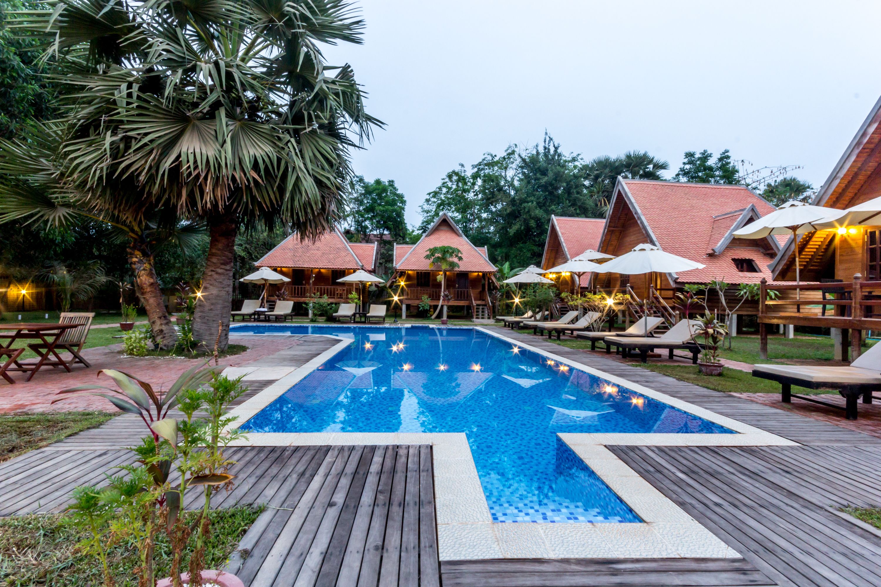 A pool area with wooden deck, lounge chairs, umbrellas, and a palm tree.