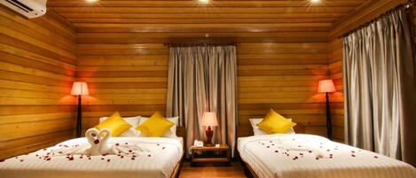 A room with two beds, wooden walls, a central nightstand with a lamp, and a ceiling with recessed lighting.