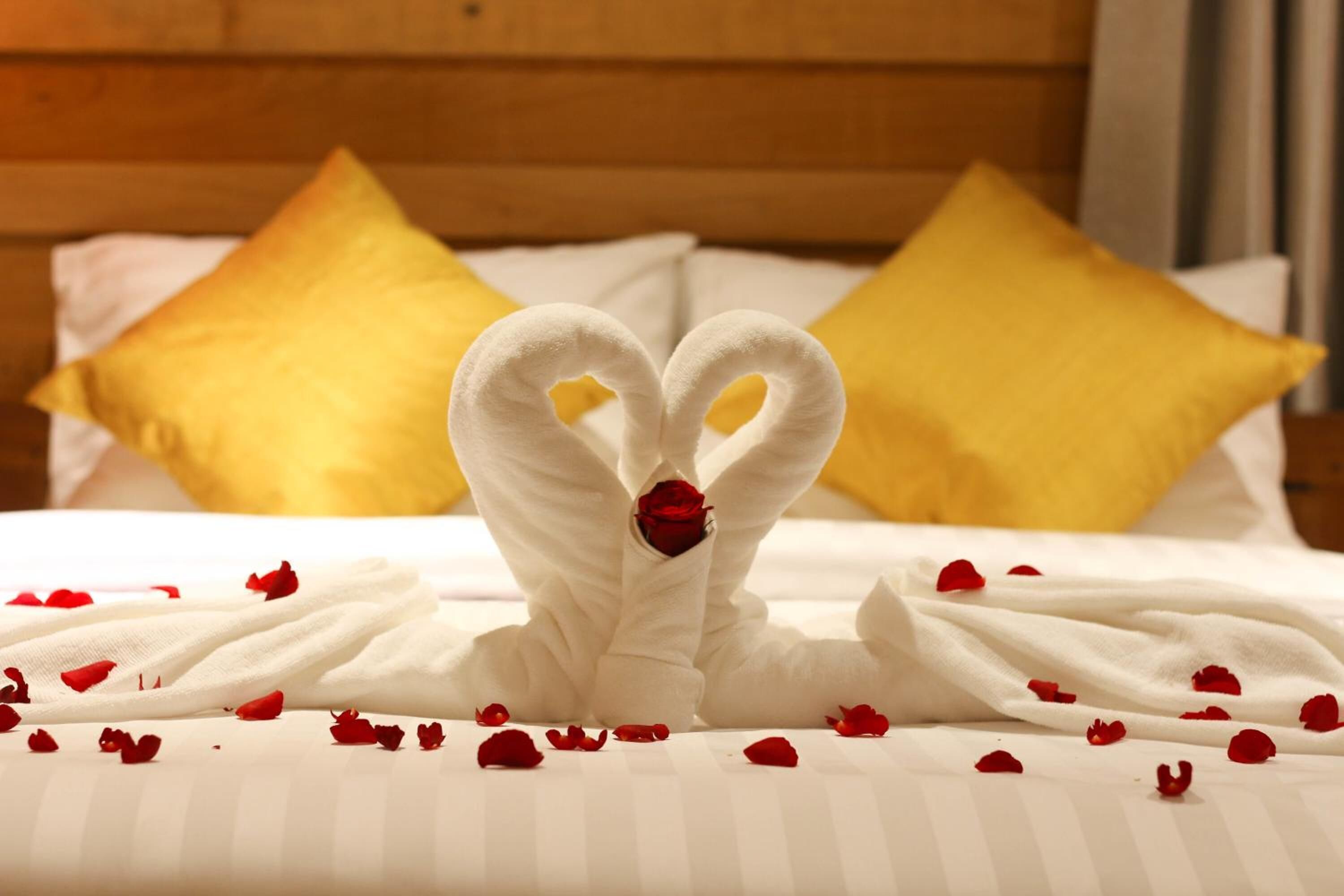 A bed with a heart-shaped towel arrangement and a rose, surrounded by scattered rose petals.