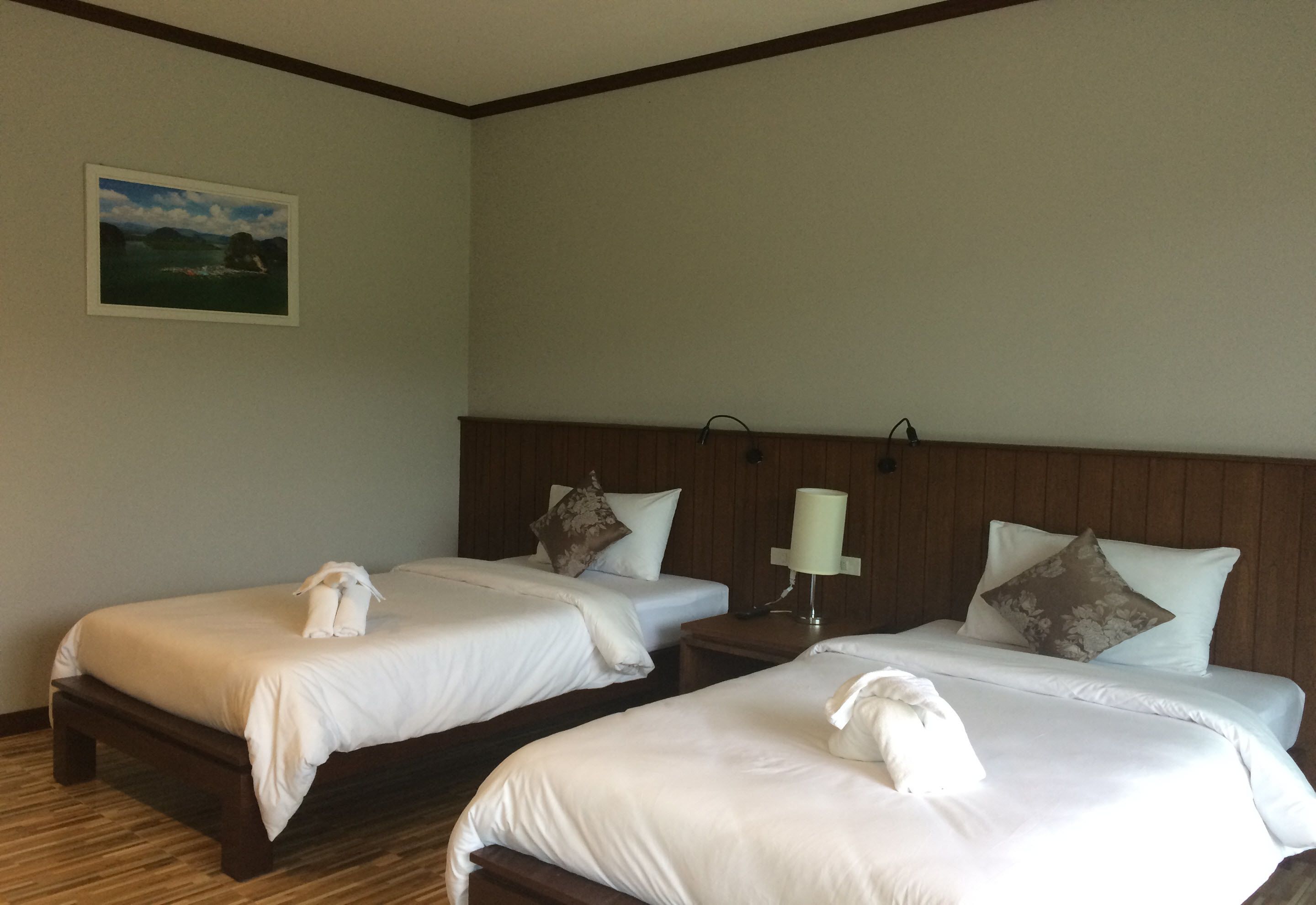 A hotel room with two single beds, wooden headboards, white linens, a bedside lamp, and a painting on the wall.