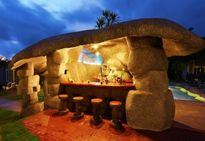 A tiki bar with stone pillars and stools, a bar counter, and a pool in the background. - Khaolak Mountain View (Takua Pa)